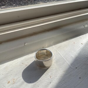 May include: A silver ring with a wide band. The ring is sitting on a white surface.