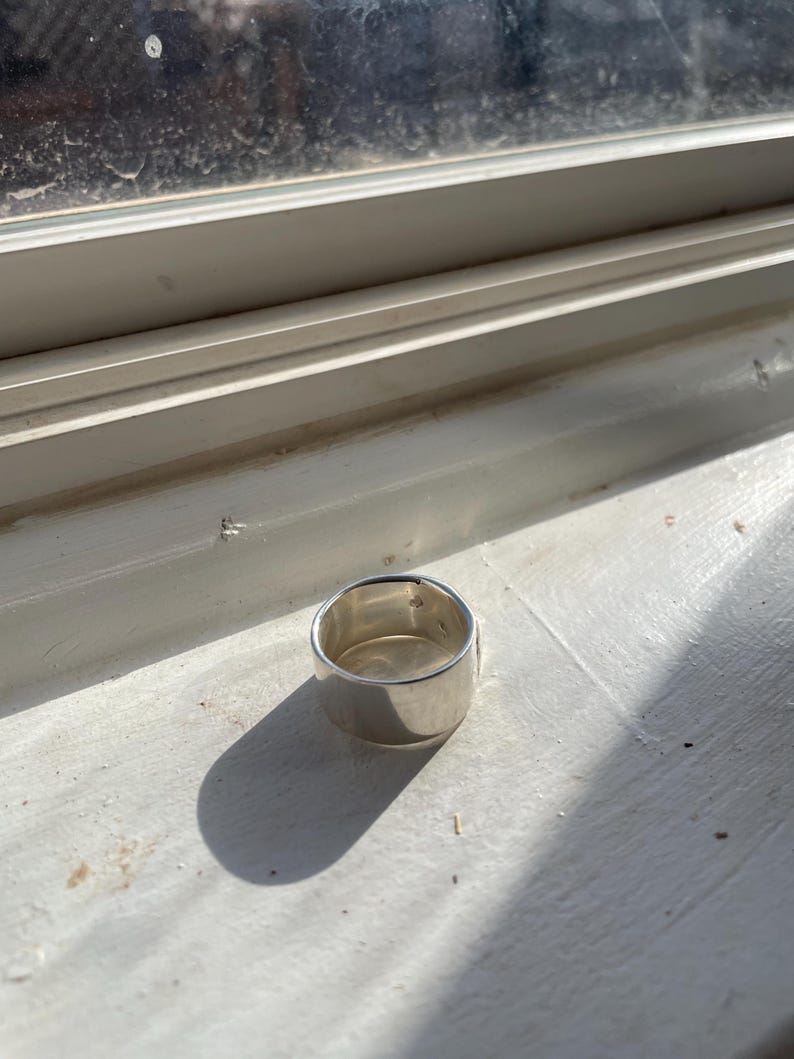May include: A silver ring with a wide band. The ring is sitting on a white surface.