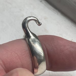 Könnte beinhalten: Ein silberner Ring mit Haken-Design. Der Ring wird an einem Finger getragen und zeigt seine gebogene Hakenform und polierte Oberfläche. Der Haken endet in einer kleinen, abgerundeten Kugel.