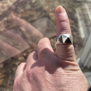 May include: A silver ring with a geometric design. The ring is worn on a finger.