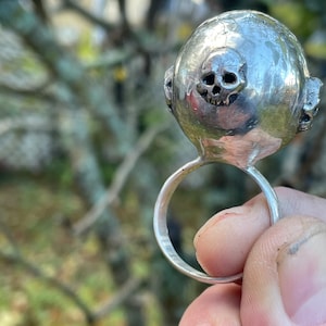 May include: A silver ring with a skull design on the top. The ring is made of metal and has a smooth finish.