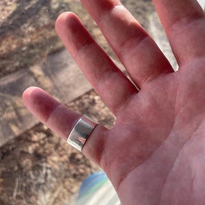 May include: A silver band ring worn on the middle finger of a hand.
