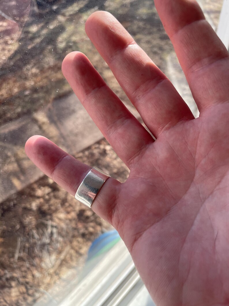 May include: A silver band ring worn on the middle finger of a hand.
