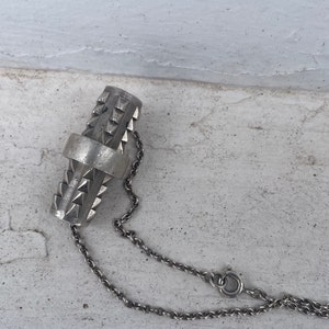 Puede incluir: Un collar de plata con un colgante cilíndrico que presenta un patrón geométrico de triángulos. El colgante está unido a una cadena de plata delicada.