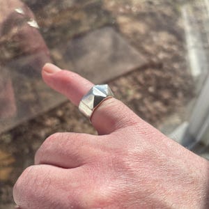 May include: A silver ring with a geometric design. The ring is worn on a finger.