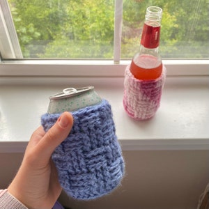 May include: Two crocheted beverage insulators, one in pink and white and one in blue. The pink and white insulator is on a glass bottle, while the blue insulator is on a can.