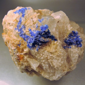 May include: A close-up of a rock with blue mineral crystals embedded in it. The rock is mostly white with some brown and orange areas. The blue crystals are clustered together in a few spots.