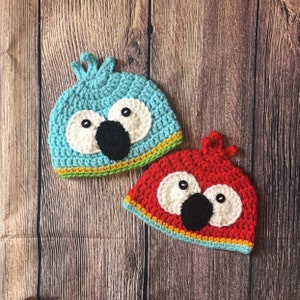 May include: Two crocheted hats with parrot faces. One hat is blue with a black beak and white eyes. The other hat is red with a black beak and white eyes. Both hats have yellow and green trim.