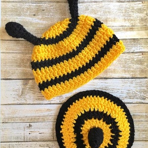 May include: A handmade crochet bee hat and matching round pad. The hat is yellow with black stripes and two black antennae. The round pad is yellow and black, with a black center. The items are made of yarn.