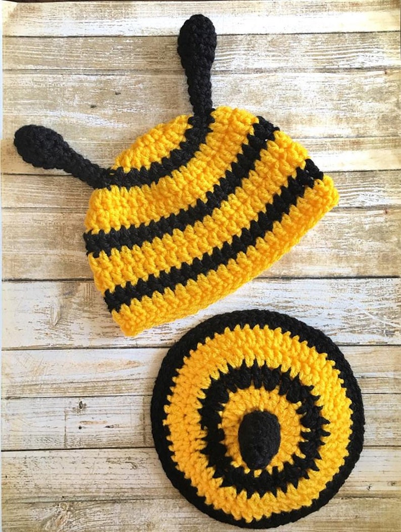May include: A handmade crochet bee hat and matching round pad. The hat is yellow with black stripes and two black antennae. The round pad is yellow and black, with a black center. The items are made of yarn.