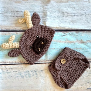 May include: A handmade crochet set featuring a brown deer hat with tan antlers and a matching diaper cover. The hat has a dark brown patch with embroidered details. The diaper cover has a wooden button closure. Perfect for baby photos.
