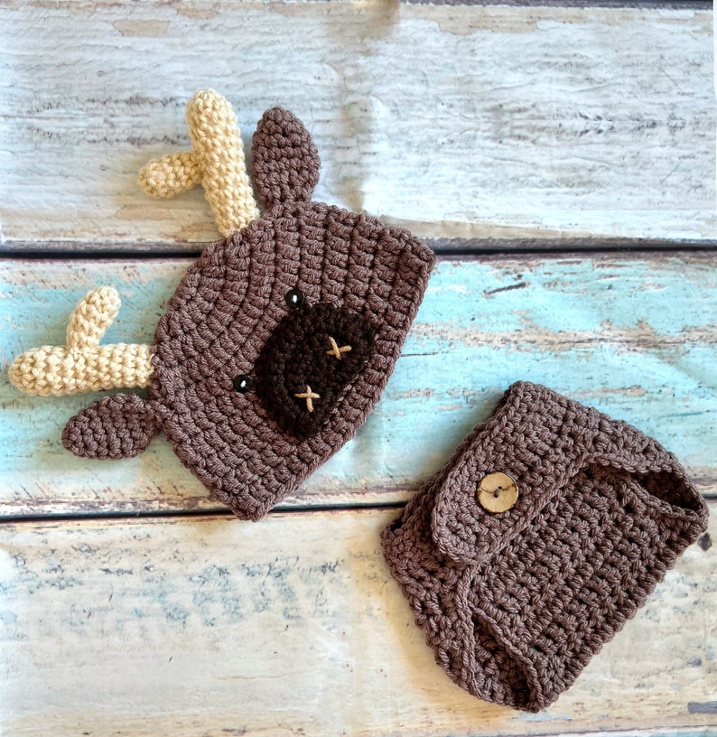May include: A handmade crochet set featuring a brown deer hat with tan antlers and a matching diaper cover. The hat has a dark brown patch with embroidered details. The diaper cover has a wooden button closure. Perfect for baby photos.
