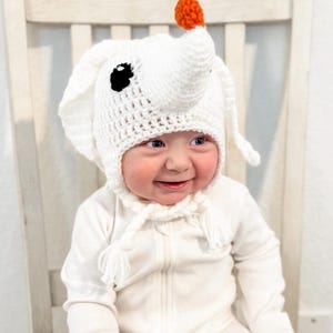 May include: A white crocheted baby hat with a dog-like design, featuring black eyes and an orange pom-pom on top. The hat is worn by a smiling baby in a white onesie. The hat has ear flaps and a tie closure.