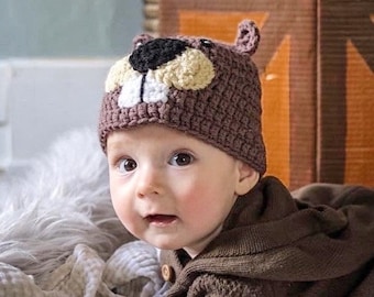 May include: A baby wearing a brown crocheted hat shaped like a beaver. The hat features a black nose, tan cheeks, white teeth, and small ears. The baby is also wearing a brown hooded jacket.