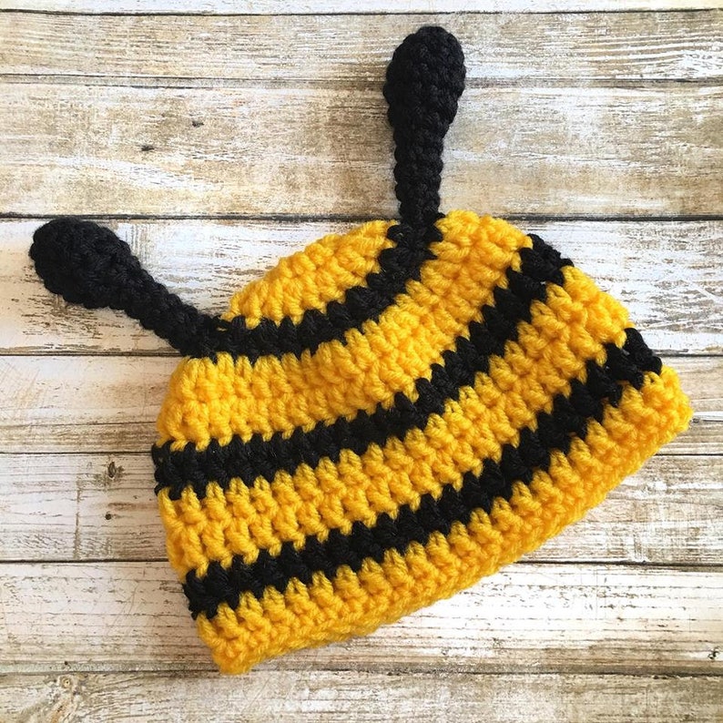 May include: A handmade crochet hat in a bee design. The hat features alternating yellow and black stripes, with two black antennae extending from the top. The hat is designed for infants and toddlers.