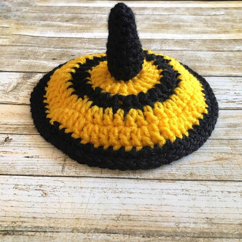 May include: A handmade crochet bee-themed hat in yellow and black. The hat is round with concentric yellow and black stripes, and a black stinger-like protrusion on top. The hat is displayed on a light wood surface.