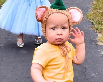 May include: A tan crocheted hat featuring a pointed green top and large, rounded ears with pink centers. The hat is designed with a chin strap that ties under the chin, providing a snug fit around the head. The texture is consistent with yarn.