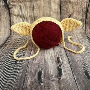 May include: A crocheted baby hat with floppy ears in a light beige color. The hat is lying on a wooden surface with a ball of maroon yarn.
