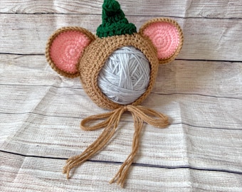 May include: A tan crochet hat featuring a pointed dark green top, two large round ears with pink centers, and long tan ties. The hat is shaped like a mouse or animal head and is shown with a ball of light gray yarn inside the face opening.