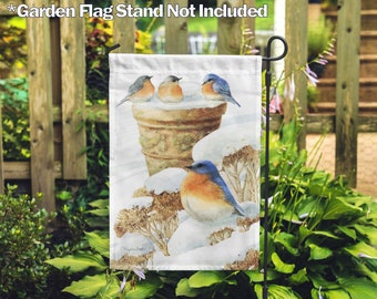 May include: A garden flag featuring a watercolor painting of bluebirds perched on a snow-covered planter and branches. The flag is displayed on a black metal stand amidst lush green foliage, creating a serene outdoor scene.