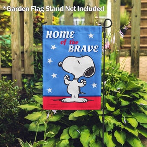Peut inclure: Un drapeau de jardin bleu avec un motif rouge, blanc et bleu représentant Snoopy debout, les pattes levées, et le texte "Home of the Brave".
