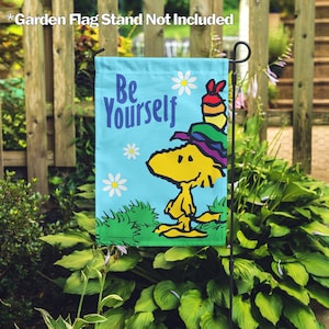 May include: A blue garden flag with a cartoon character wearing a rainbow hat and the text "Be Yourself". The flag is displayed on a black metal stand in a garden setting.