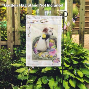 May include: A white dog with brown ears sits in a wicker basket filled with colorful flowers. The dog is wearing a black bow tie and a yellow flower behind its ear. The image is a garden flag with the text "Magnolia Garden" in the bottom right corner.