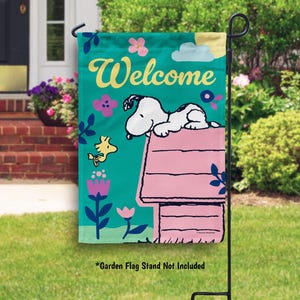 May include: A green garden flag with a pink dog house and a white dog with black spots on top of the house. The dog is looking to the left. The flag has the word "Welcome" in yellow letters and a yellow bird flying to the left. The flag has a green background with pink, yellow, and purple flowers.