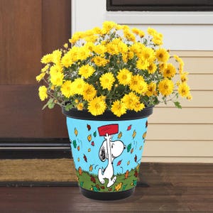 May include: A black and blue Snoopy flower pot filled with bright yellow chrysanthemums. The pot features Snoopy holding a red sign, with falling leaves on a blue background. The pot is sitting on a wooden surface.