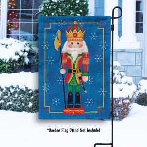 May include: A blue garden flag featuring a colorful illustration of a nutcracker soldier. The nutcracker wears a red and green uniform, a gold crown, and holds an axe. White snowflakes decorate the blue background. The flag is displayed on a black garden flag stand.