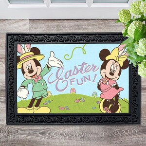 Op de afbeelding: Een zwarte deurmat met een witte rand en een kleurrijk ontwerp met Mickey en Minnie Mouse gekleed in paasoutfits. De tekst "Easter Fun!" is in paars op een blauwe achtergrond geschreven.