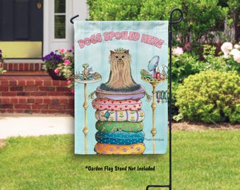 May include: A garden flag with the text "DOGS SPOILED HERE" features a pampered dog atop a stack of colorful dog beds. The flag has a light blue background and is displayed on a black metal stand in a garden setting.