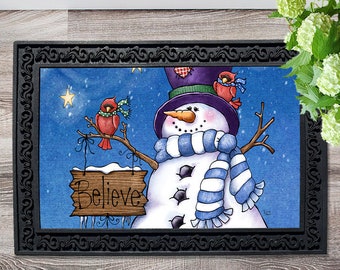 May include: A rectangular doormat with a black ornate border. The mat features a snowman with a purple top hat, blue and white scarf, and a carrot nose. Two red cardinals perch on branches. A sign that says "Believe" hangs from the branches.