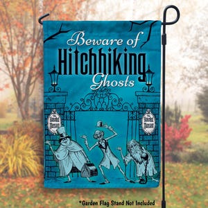 May include: A blue and white garden flag with the text "Beware of Hitchhiking Ghosts" and an image of three ghosts from the Haunted Mansion attraction. The flag is hanging from a black metal garden flag stand.