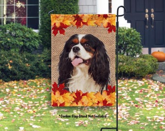 May include: A garden flag featuring a Cavalier King Charles Spaniel with a burlap background and a border of autumn leaves. The dog has black, white, and brown fur and a pink tongue. The flag is displayed on a black metal stand.