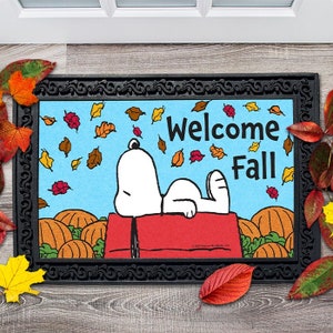 May include: A welcome mat with a black border and a blue background featuring Snoopy laying on a red doghouse with pumpkins and falling leaves. The text "Welcome Fall" is written in white letters.