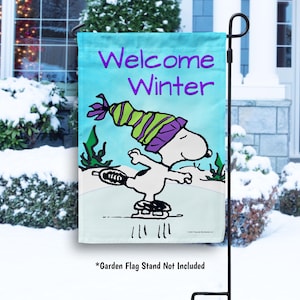 May include: A garden flag with a blue background featuring a cartoon character ice skating. The character is wearing a green and purple hat. The text "Welcome Winter" is printed in purple on the flag.