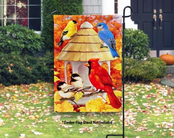 May include: A decorative garden flag featuring a birdhouse with several colorful birds. A yellow bird perches on the roof, a blue bird sits on the side, and a red cardinal is perched below. The background is a fall-themed scene.