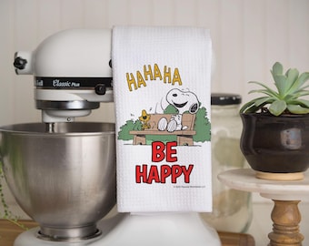 May include: White kitchen towel with a cartoon image of Snoopy and Woodstock. The text on the towel reads "HAHAHA BE HAPPY".