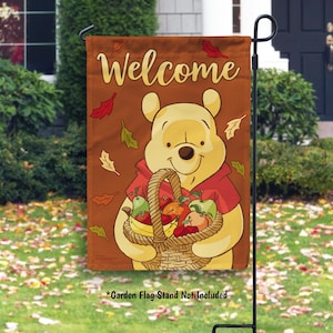 May include: A garden flag featuring a cartoon bear holding a basket of fruit. The flag has the word "Welcome" at the top and is decorated with autumn leaves. The background is a brown color.