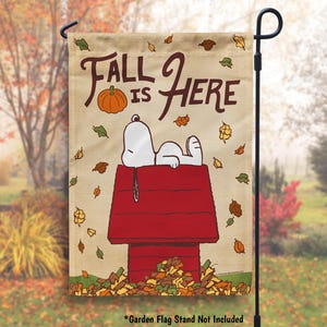 May include: A beige garden flag with a cartoon dog lying on a red doghouse. The flag has the text "Fall is Here" with a pumpkin and falling leaves. The text "Garden Flag Stand Not Included" is at the bottom.