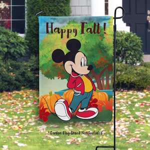 May include: A garden flag featuring Mickey Mouse in a red jacket, yellow shirt, and blue pants, with pumpkins and fall foliage. The flag reads "Happy Fall!" in brown letters. The garden flag is displayed on a black metal stand.