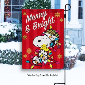 May include: A red garden flag with the words "Merry & Bright" and a cartoon image of Snoopy and Woodstock wrapped in Christmas lights. The flag has a vertical striped pattern and snowflake accents.