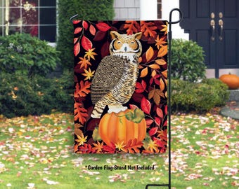 May include: A decorative garden flag featuring a detailed illustration of a great horned owl perched on an orange pumpkin. The background is a dark color with autumn leaves in shades of red, orange, and yellow. The text "*Garden Flag Stand Not Included" is at the bottom.