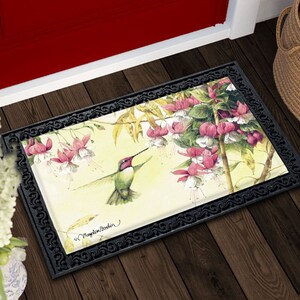 Op de afbeelding: Rechthoekige deurmat met een zwarte decoratieve rand. De mat heeft een aquarel van een kolibrie met een lange snavel, zwevend in de buurt van roze en witte bloemen. De achtergrond is zachtgeel.