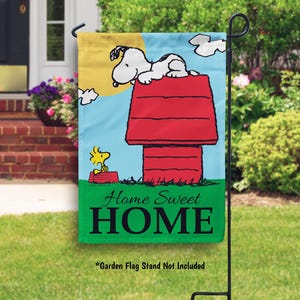May include: A colorful garden flag featuring Snoopy and Woodstock from Peanuts. Snoopy is on top of a red doghouse with the text "Home Sweet HOME".