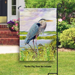 May include: A garden flag featuring a watercolor painting of a blue heron standing in a marsh. The heron is blue-grey with a long neck and long legs. The background is a soft blue and green with a hint of purple.