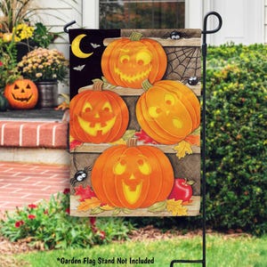 May include: A Halloween garden flag featuring five carved orange pumpkins with smiling faces. The flag has a black background with a yellow crescent moon, bats, spiders, and a spider web design. The flag is displayed on a black metal stand.