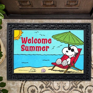 May include: A black doormat with a white border and a cartoon image of Snoopy relaxing on a beach chair under an umbrella. The text "Welcome Summer" is printed in red on the doormat.