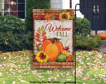 May include: A decorative garden flag with a fall theme. The flag features the words "Welcome Fall" above a design of pumpkins, sunflowers, and autumn leaves. The background has a rustic wood and plaid pattern.